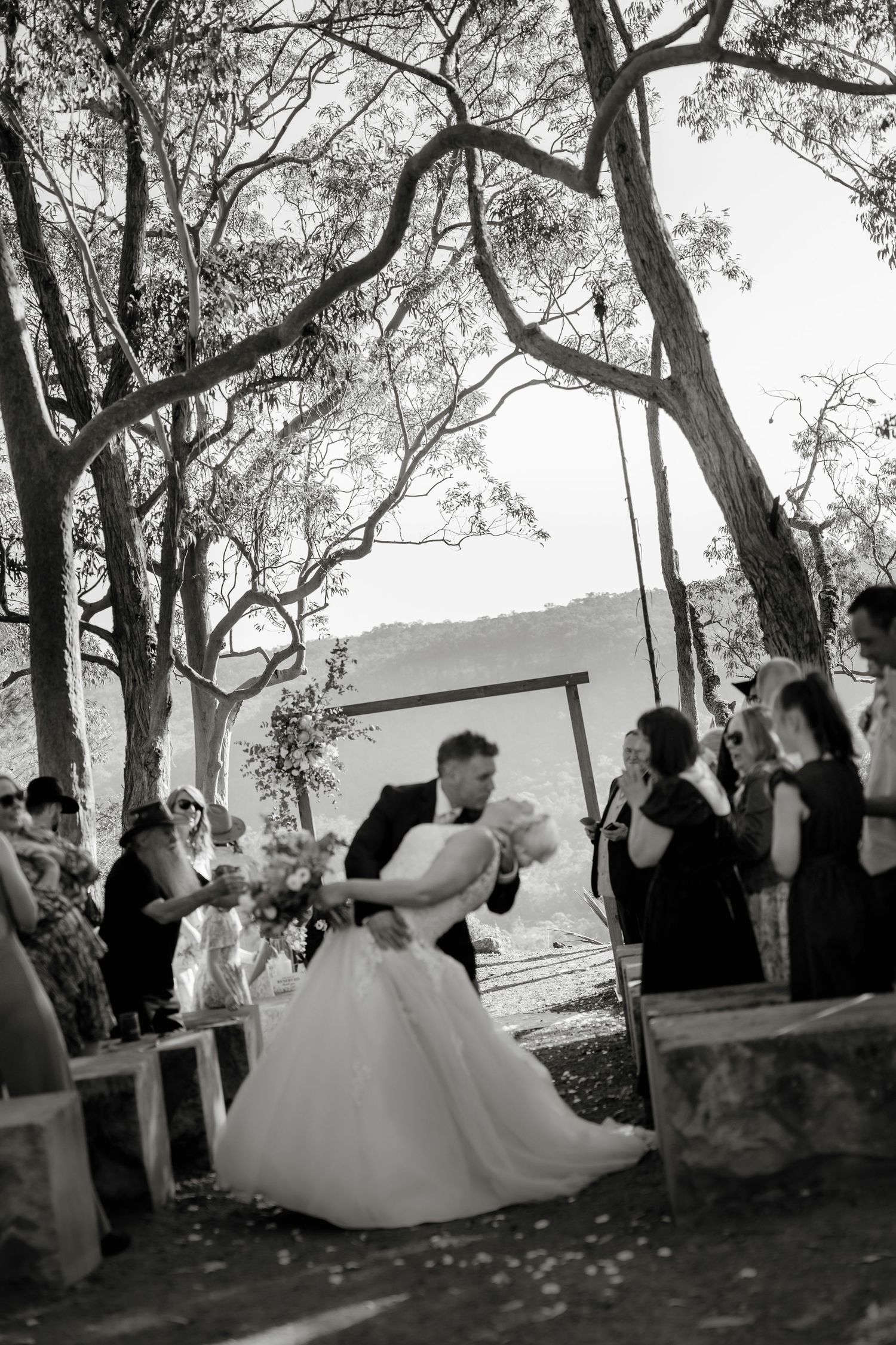 Black and white photo of bride and groom sharing first kiss at outdoor wedding ceremony under tall trees.