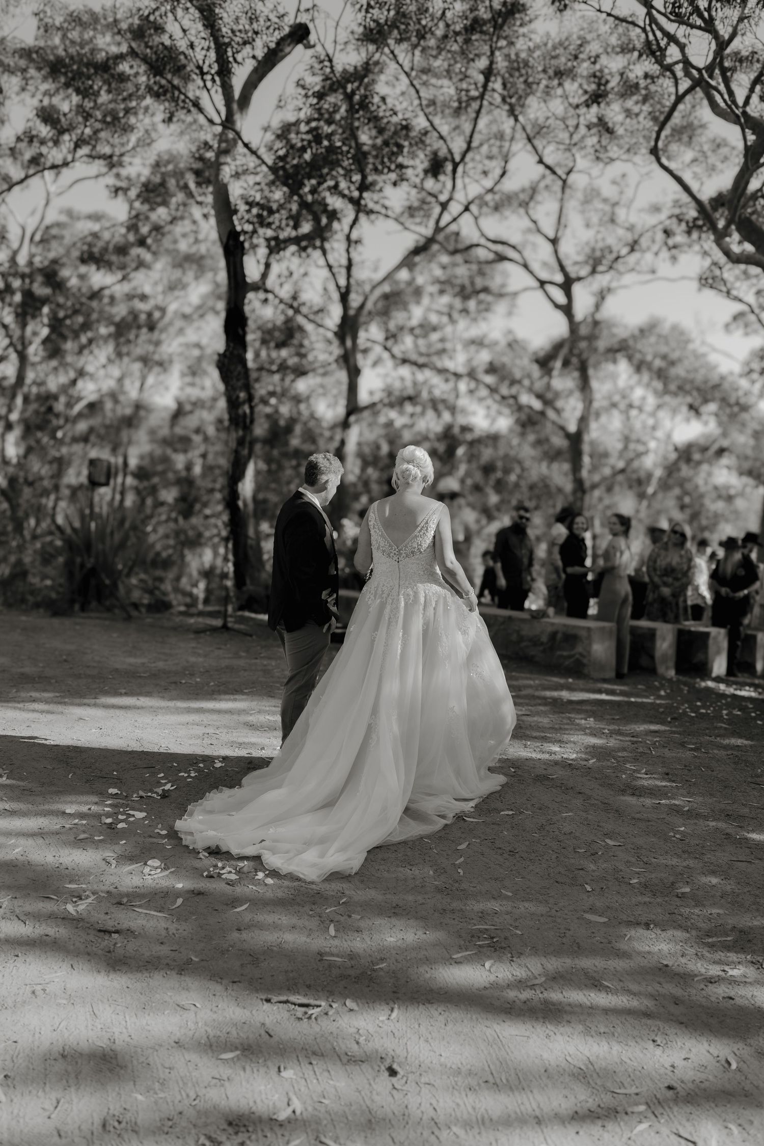 Black and white photo of a bride in a flowing gown walking with groom toward wedding guests outdoors.