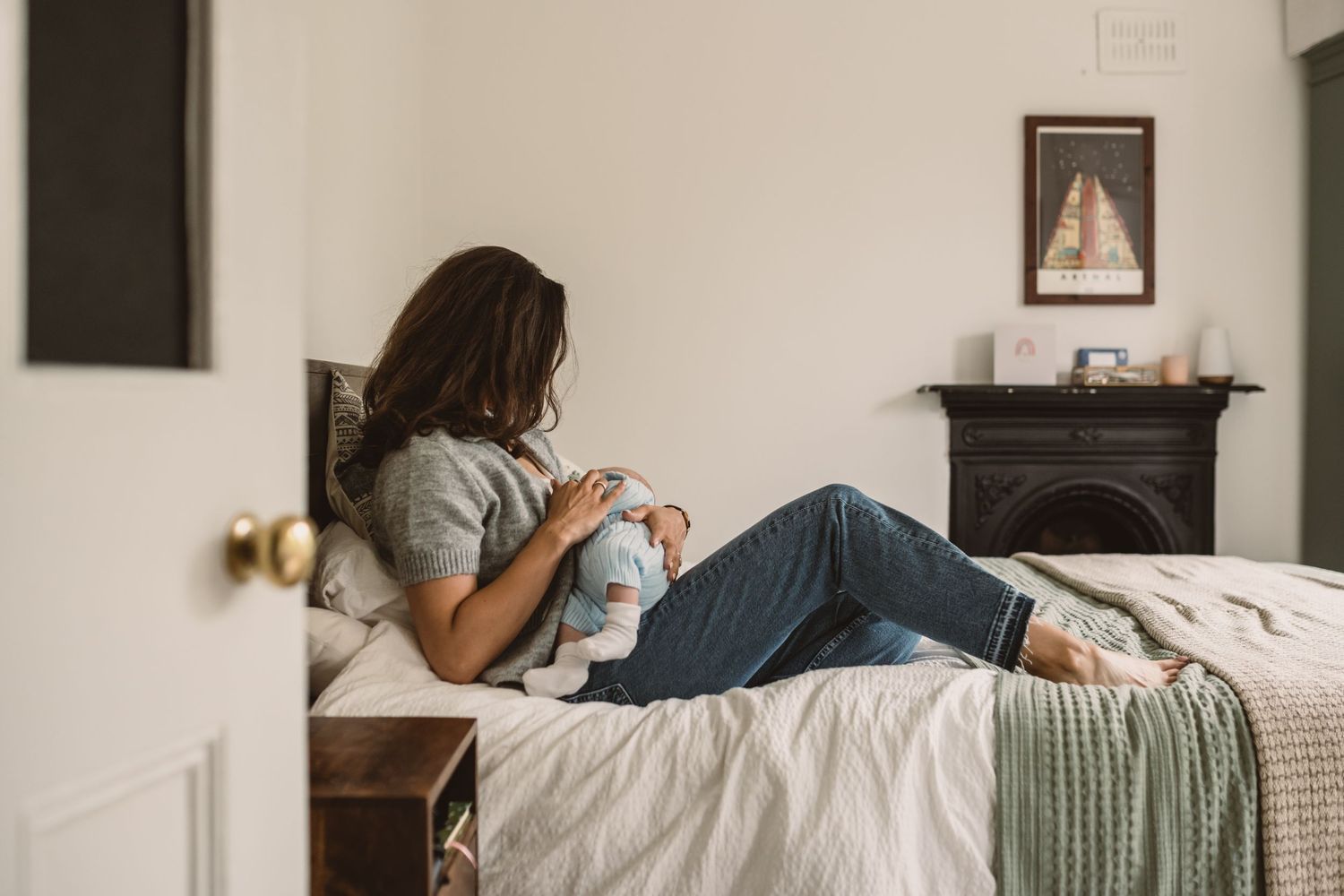 Mother sitting on bed breastfeeding her newborn baby in a cozy bedroom with fireplace.