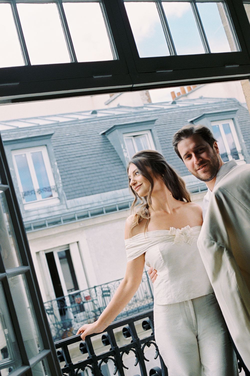 Elegant couple in white attire standing on a Parisian balcony with classic Haussmann architecture in the background. civil wedding