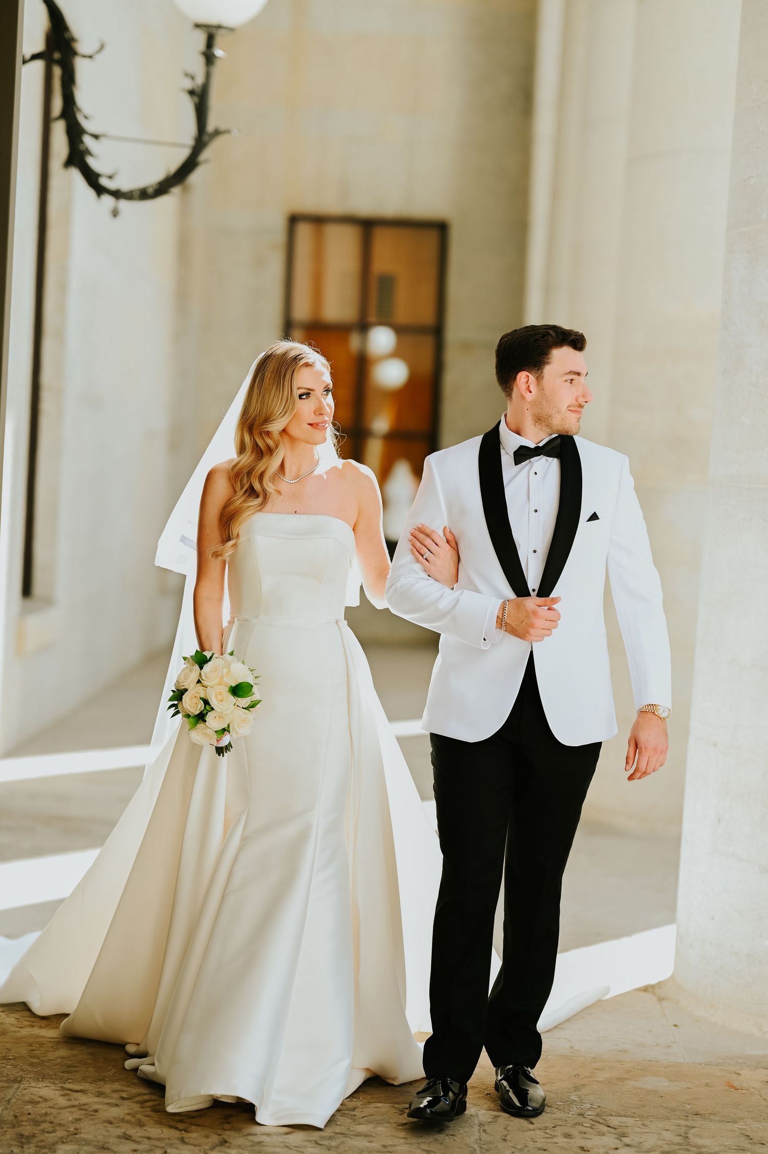 Bride in strapless white gown and groom in white tuxedo jacket walking together under stone archway.