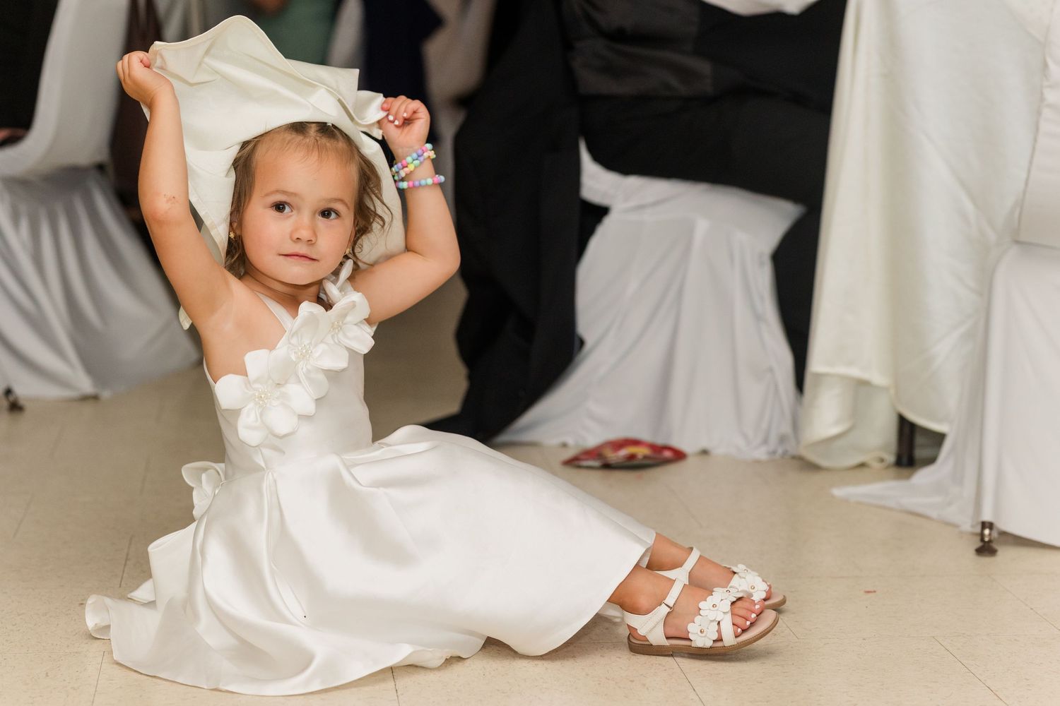 Young flower girl in white dress sits on floor holding fabric above her head at a wedding reception.