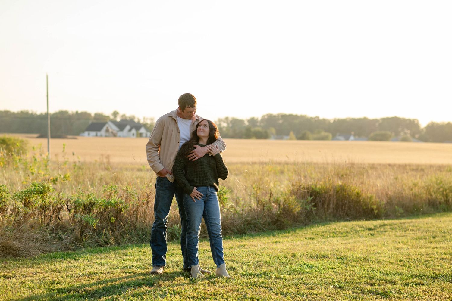 Smiling couple embraces in golden sunlit field during outdoor engagement photo session at sunset.