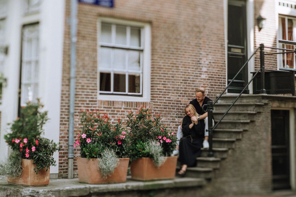 Couple embracing on brick building steps surrounded by colorful potted flowers in a charming Amsterdam street setting.