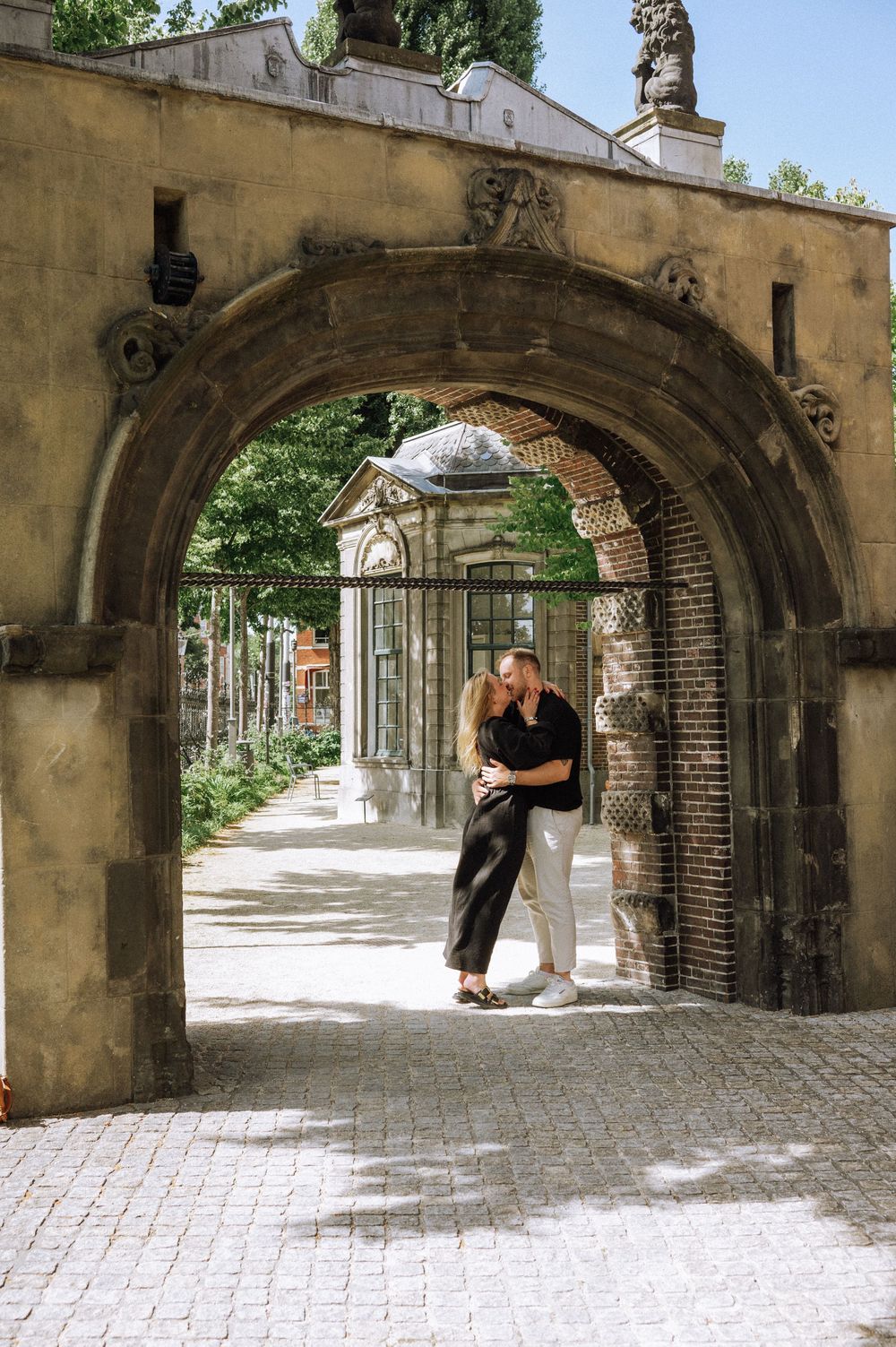 Couple kissing under a historic stone archway in Rijksmuseum with ornate details and lush greenery in the background.