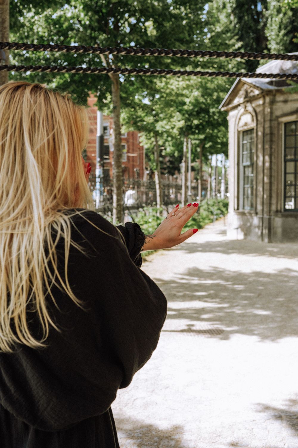 Blonde woman in black jacket gesturing with red nails near a fence in a tree-lined courtyard.