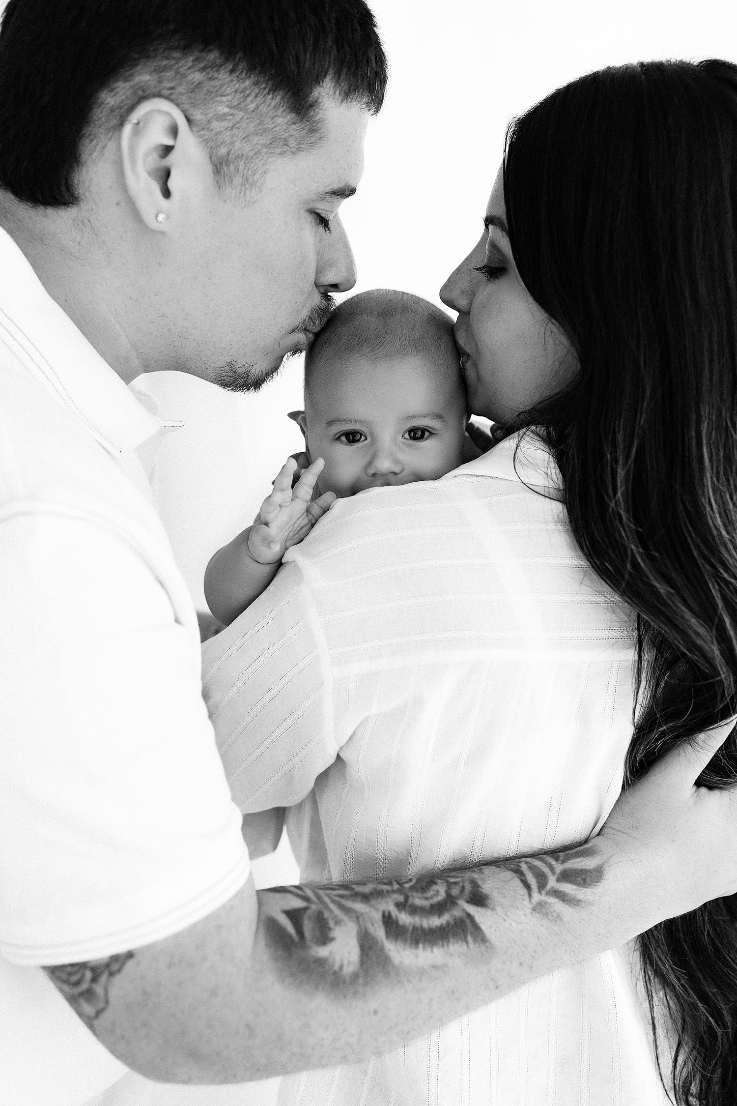 Black and white photo of parents kissing their newborn baby, father with tattoos, all dressed in white.