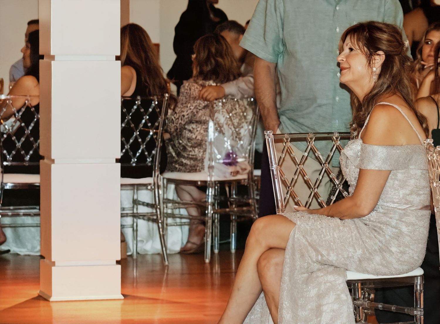 Smiling woman in a shimmering silver dress sits beside a clear acrylic chair at an indoor event with guests seated behind her.