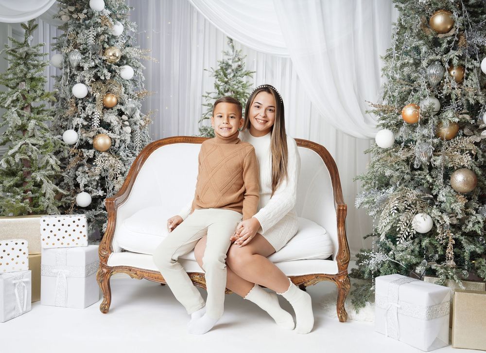 Mother and son in cream outfits sit on a vintage white sofa surrounded by frosted Christmas trees, gold ornaments, and wrapped gifts.