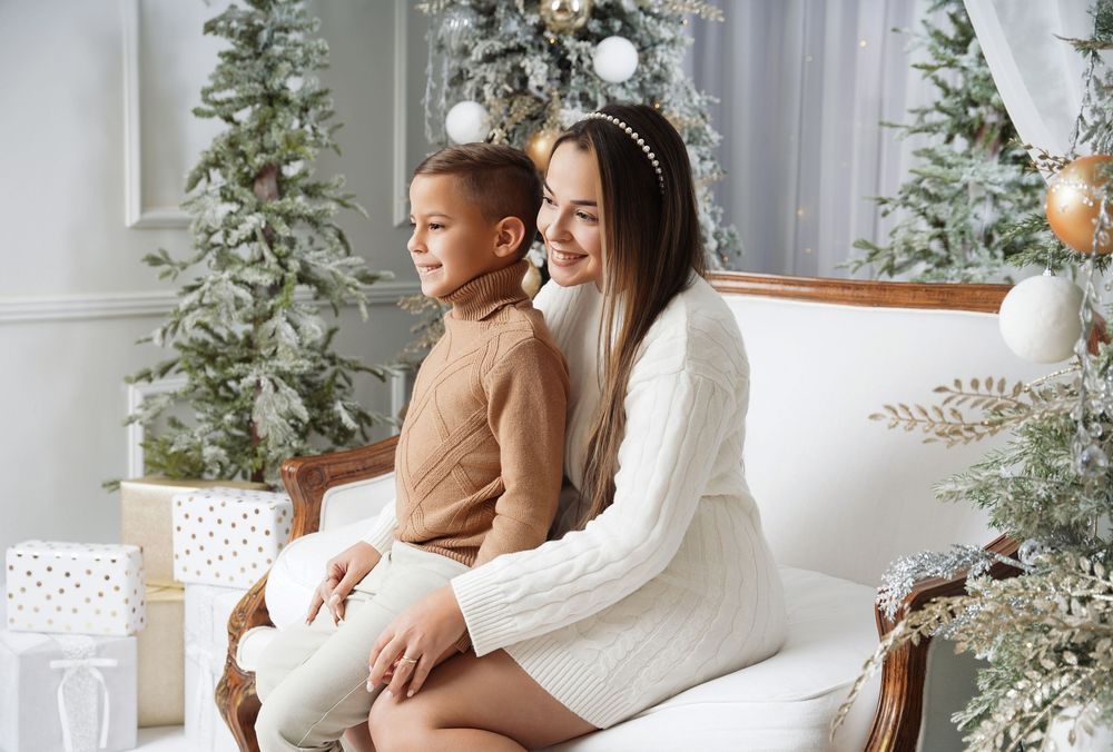 Smiling mother in a cream knit dress sits with her young son on a white sofa beside decorated Christmas trees and wrapped gifts.