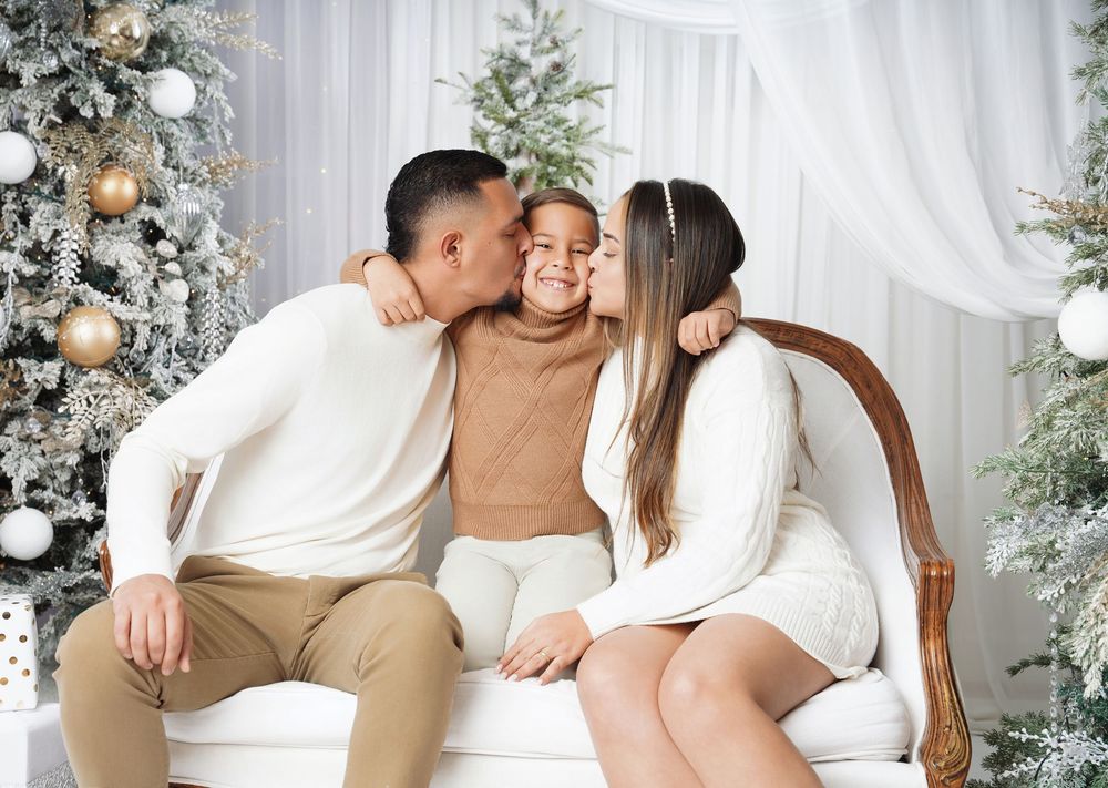 Family in cream and tan outfits sharing a kiss with a smiling child on a vintage bench beside decorated Christmas trees in a studio.
