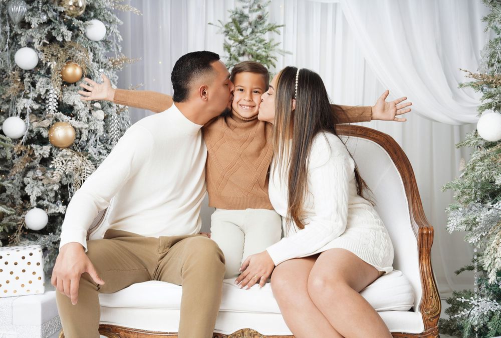 Man and woman kiss a smiling child's cheeks on a cream sofa beside a decorated white Christmas tree in a festive family holiday portrait.