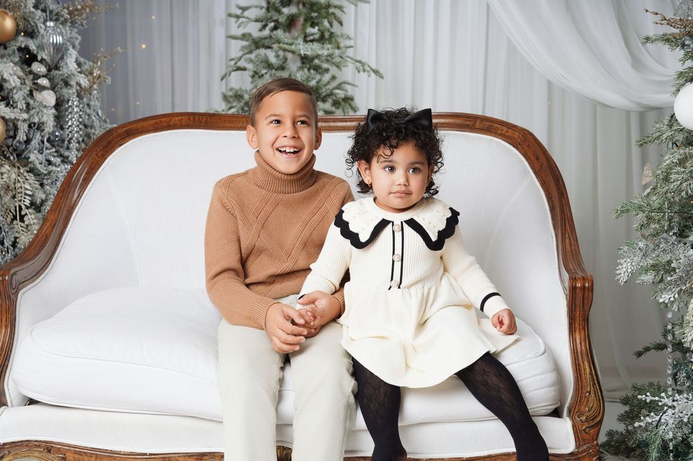 Two smiling children in cozy neutral outfits sit together on a white vintage sofa in a festive Christmas photo studio with decorated trees.