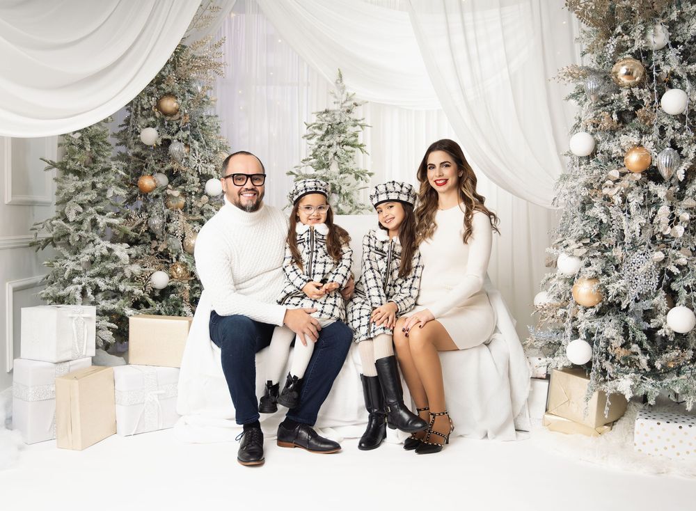 Family of four in matching white and plaid outfits posing on a white set surrounded by frosted Christmas trees and gold ornaments.