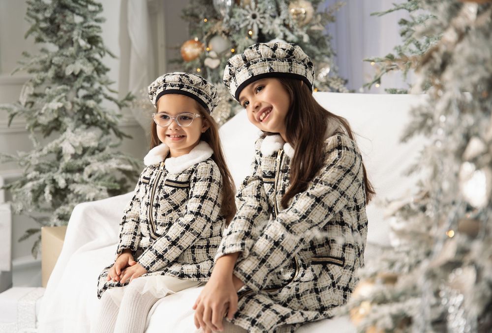 Two smiling girls in matching black and white tweed jackets and berets sit together beside frosted Christmas trees with gold ornaments.