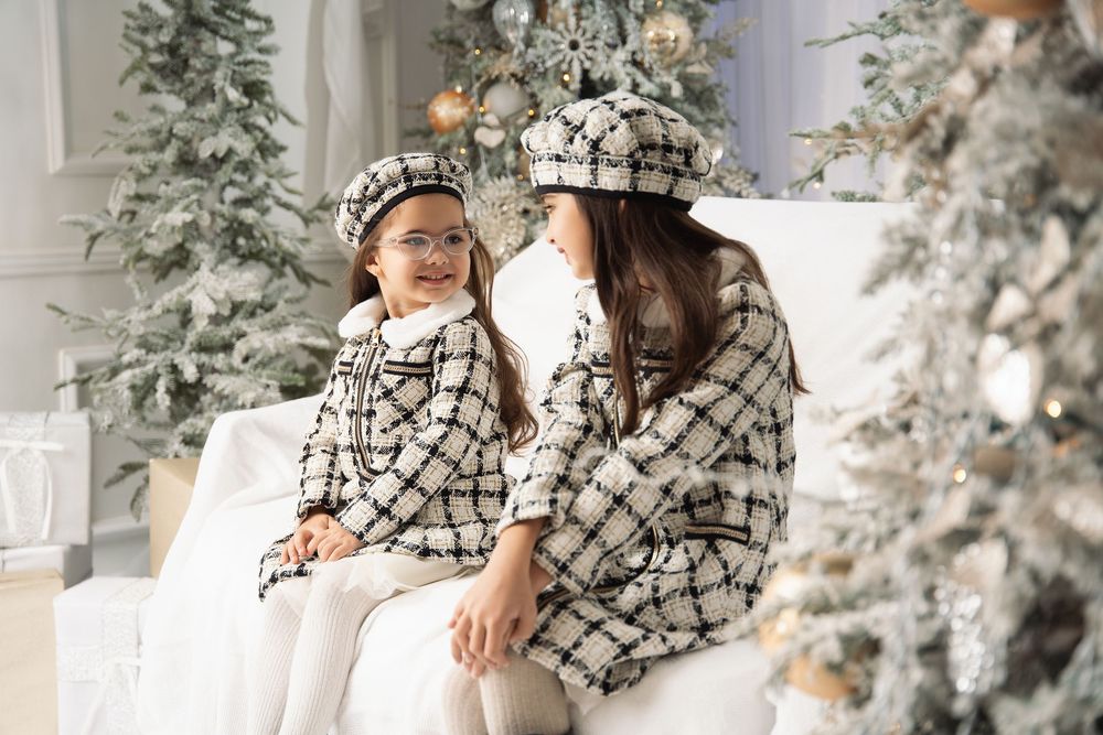 Mother and daughter in matching black and white tweed jackets and berets sit on a white sofa beside frosted Christmas trees.