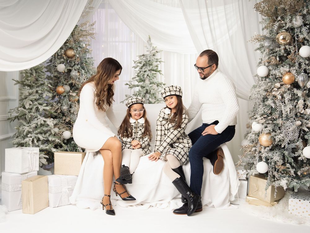 Family of four in cozy white and plaid outfits posing on a white sofa surrounded by frosted Christmas trees, ornaments, and gift boxes.