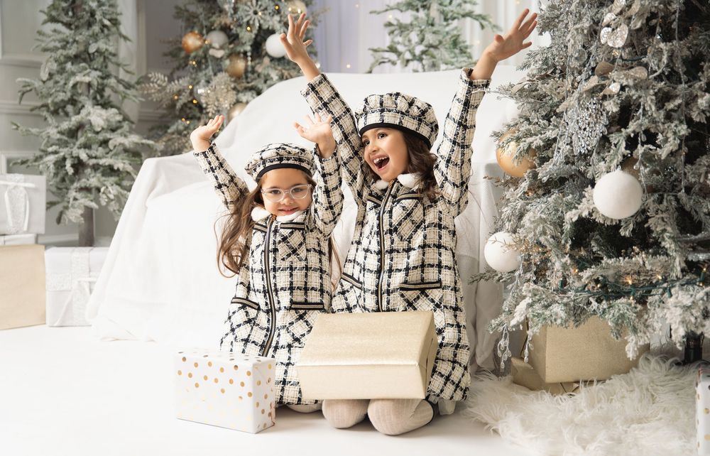 Two girls in matching black and white tweed outfits and berets cheer with raised arms beside a frosted Christmas tree and gift boxes.