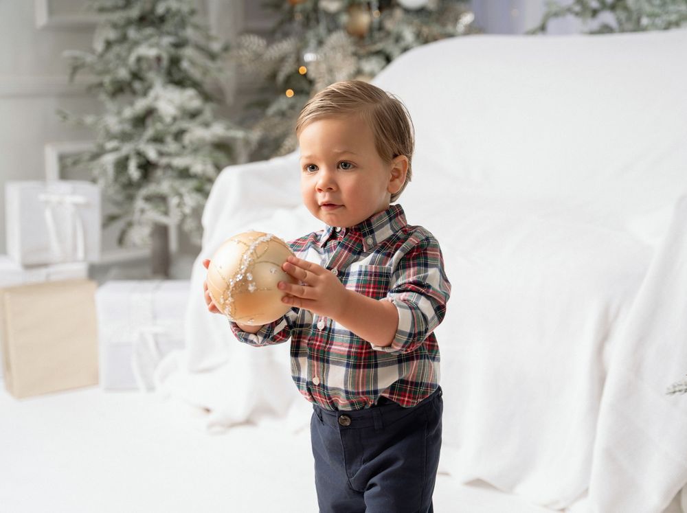 Toddler in a plaid flannel shirt holding a large gold Christmas ornament beside a white sofa and a snowy flocked holiday tree.