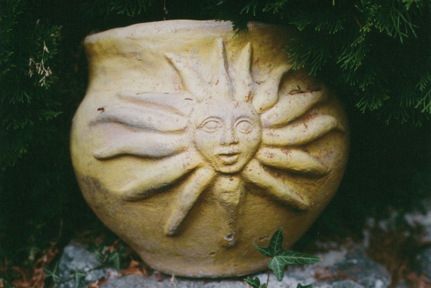 Handcrafted terracotta garden pot with a sculpted smiling sun face and radiating rays, set among green foliage and stones.