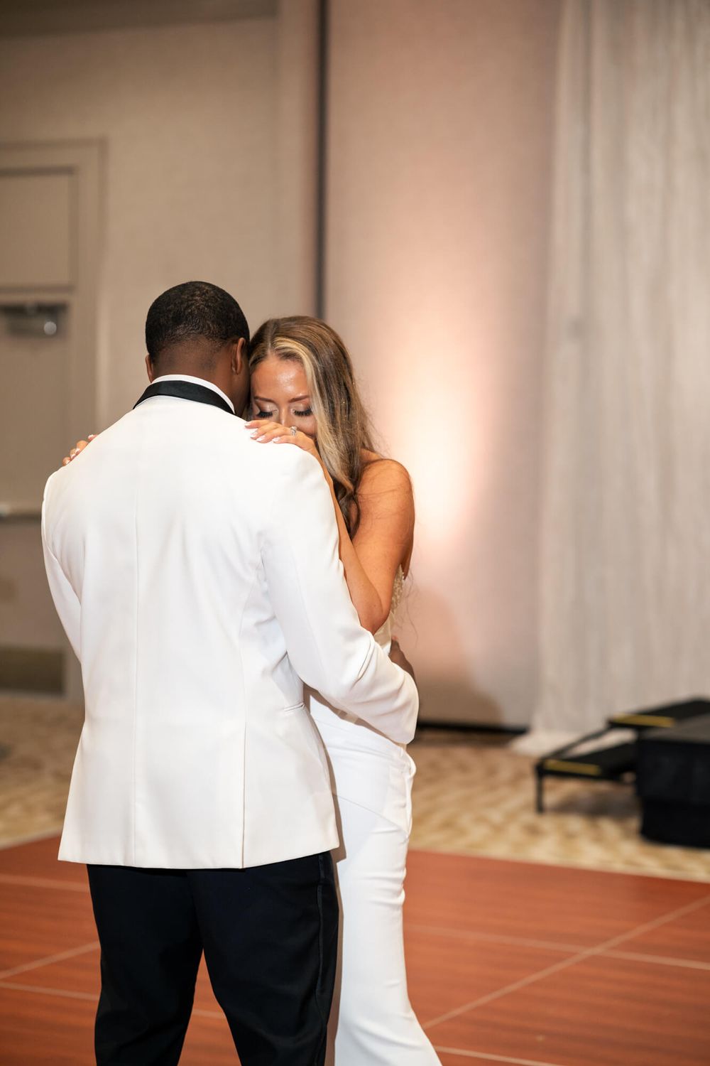 A bride in a white gown and a groom in a white tuxedo jacket share an emotional first dance on a wood dance floor at a wedding reception.