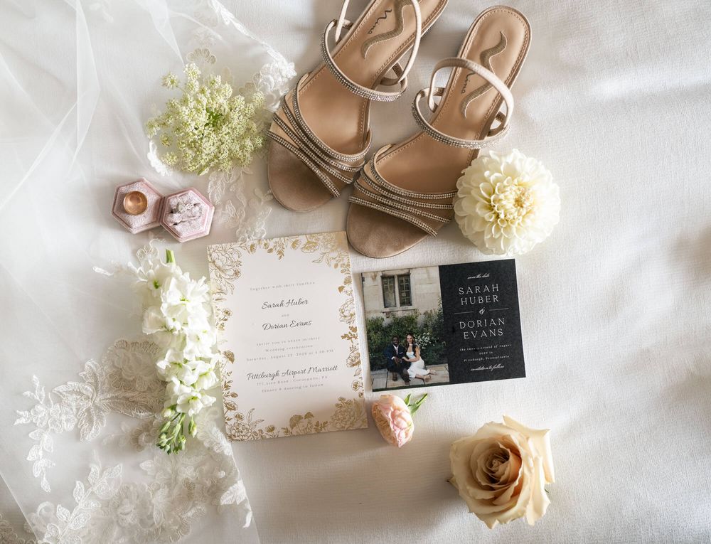 Flat lay of wedding details with tan strappy heels, floral invitation, ring boxes, cream roses and white blooms on a soft veil.