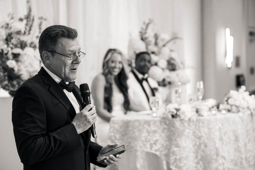 Man in a tuxedo speaks into a microphone while a smiling bride and groom listen at a flower-decorated wedding reception table.
