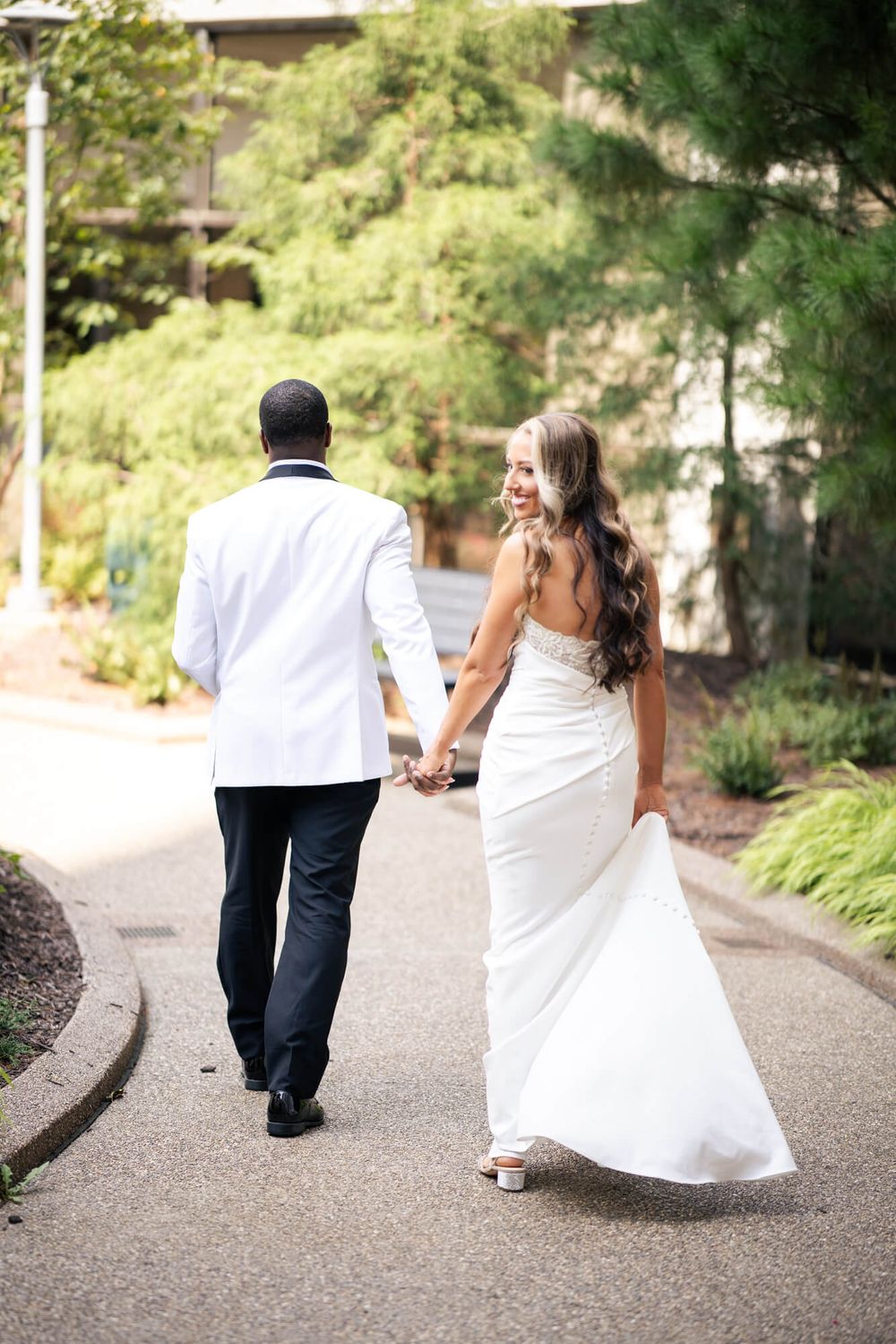 Bride in a flowing white gown holds hands with groom in a white tuxedo jacket as they walk down a garden path.