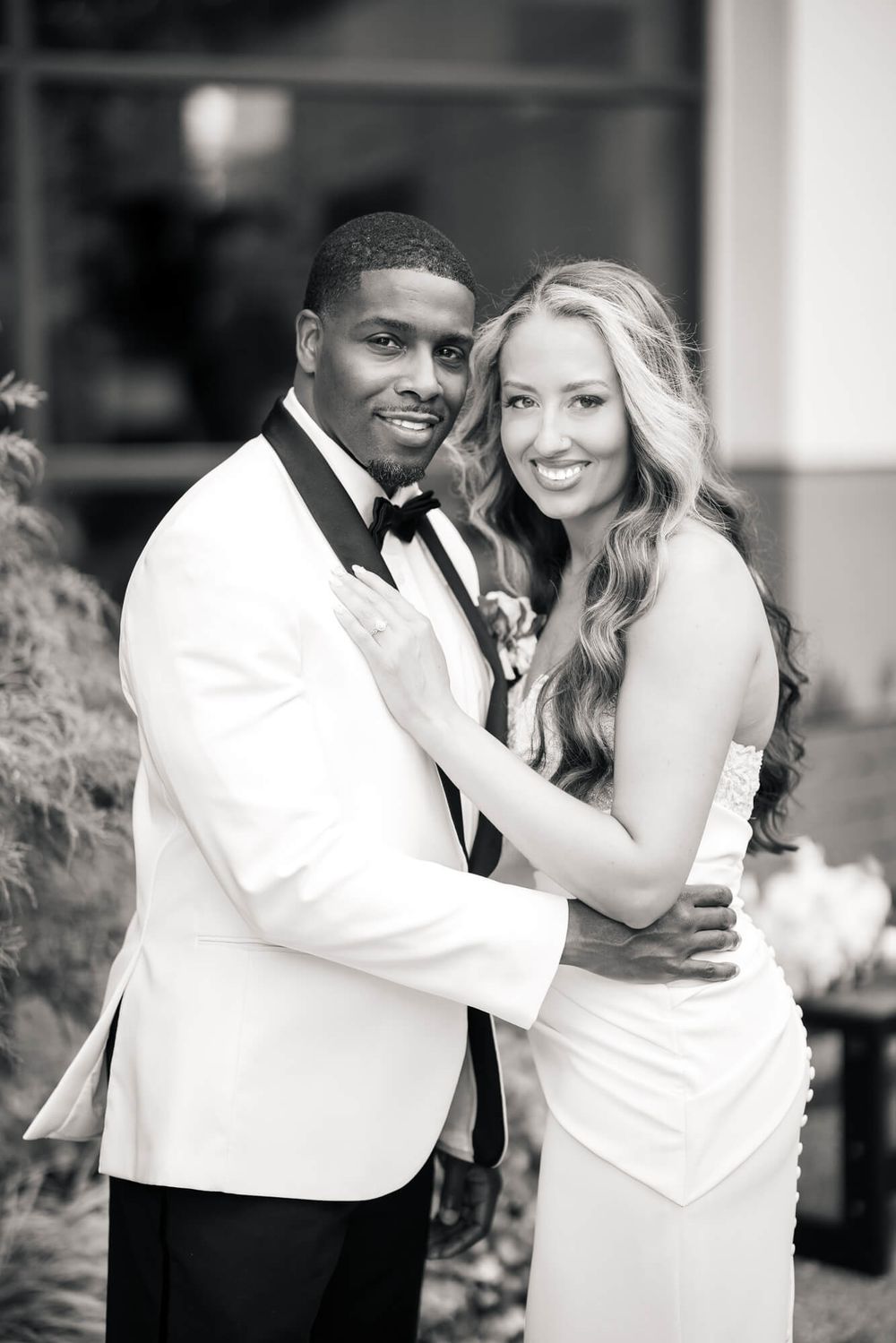 Black and white wedding portrait of a smiling bride in a strapless gown embracing her groom in a white tuxedo jacket and bow tie.
