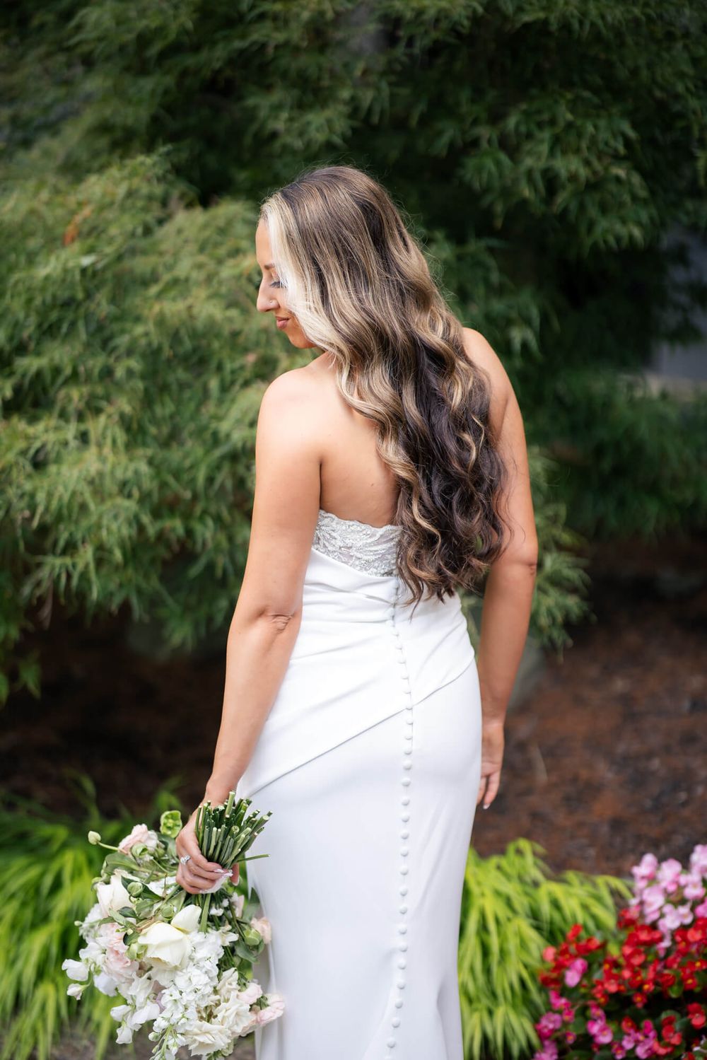 Bride in a strapless white wedding gown with button back and wavy highlighted hair holds a white bouquet in a lush garden.