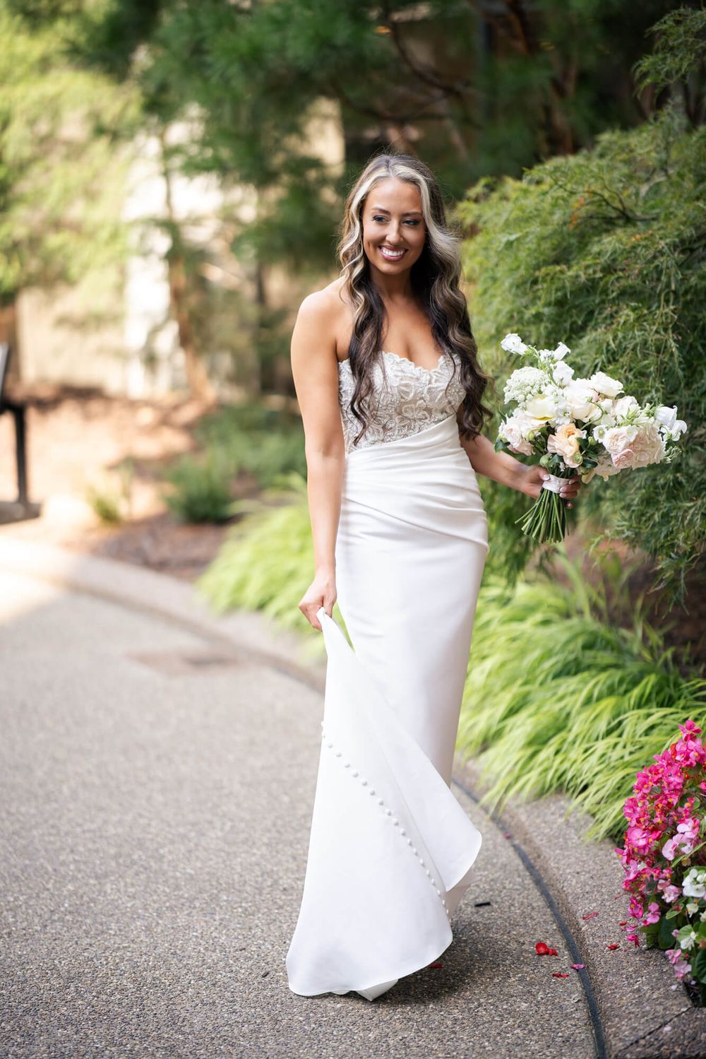 Smiling bride in a fitted white strapless lace-bodice wedding gown holds a white and blush bouquet on a garden path surrounded by greenery.