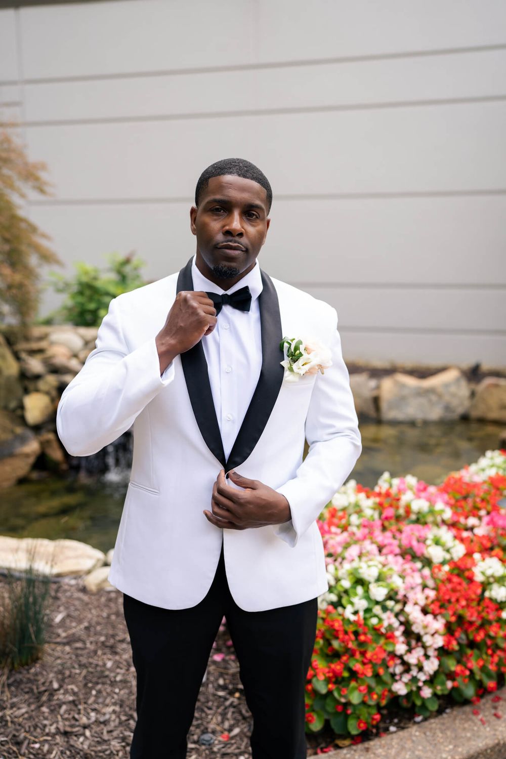 Groom in a white tuxedo jacket with black lapels and bow tie adjusts his jacket beside a flower bed and stone water feature.