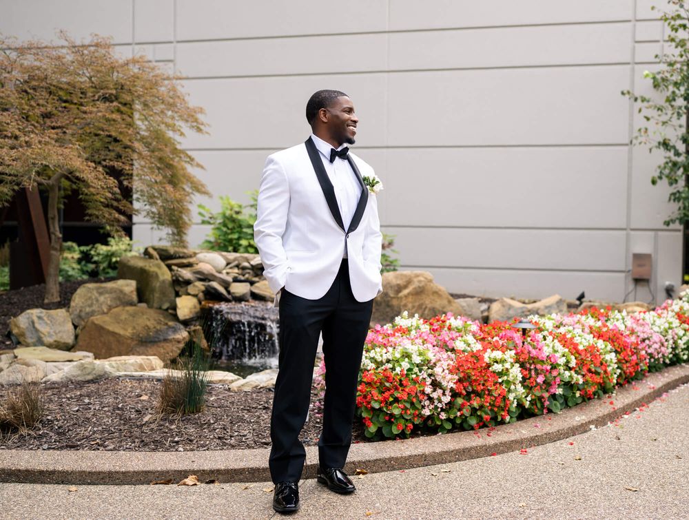 Groom in a white tuxedo jacket and black bow tie stands beside colorful flower beds and a rock water feature on a landscaped garden path.