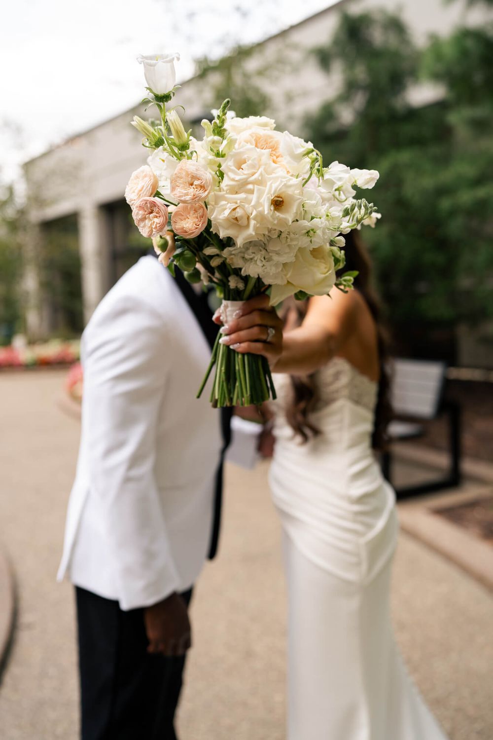 Bride and groom hide their faces behind a blush and ivory rose wedding bouquet, he in a white tuxedo jacket and she in a strapless gown.