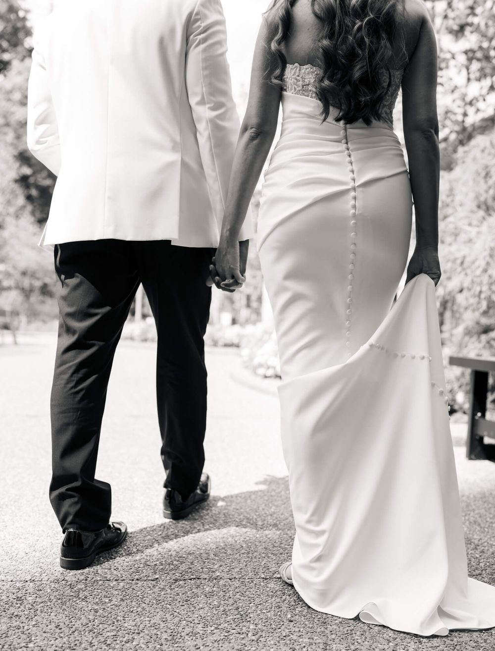 Bride in a fitted white button-back gown and groom in a white jacket hold hands while walking away in a black and white wedding photo.