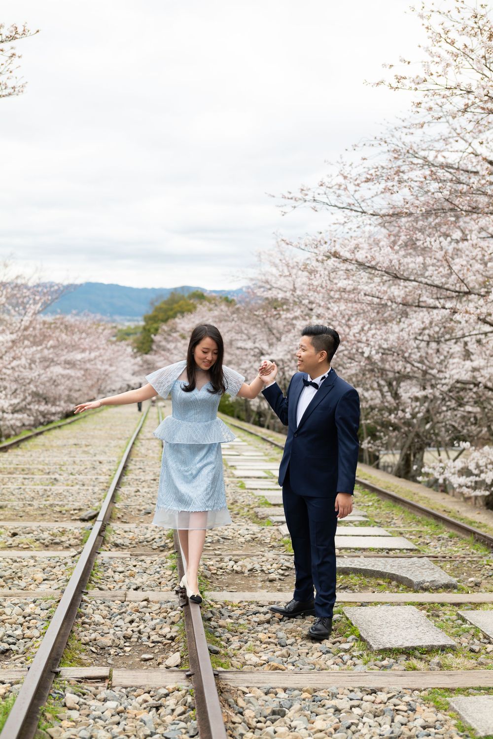 Couple in a blue dress and navy suit hold hands on old railway tracks lined with blooming cherry blossom trees.