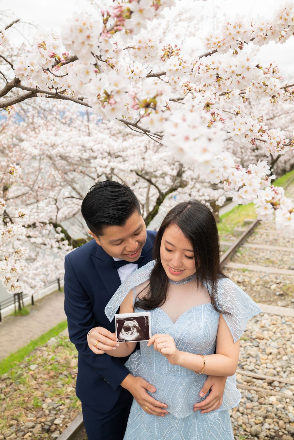 Expecting couple in a navy suit and light blue gown hold an ultrasound photo beneath blooming cherry blossoms in a maternity photoshoot.