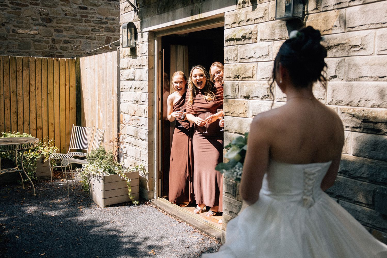 Bride in a white gown holding a bouquet stands beside a vintage cream wedding car while a chauffeur waits on a gravel driveway.