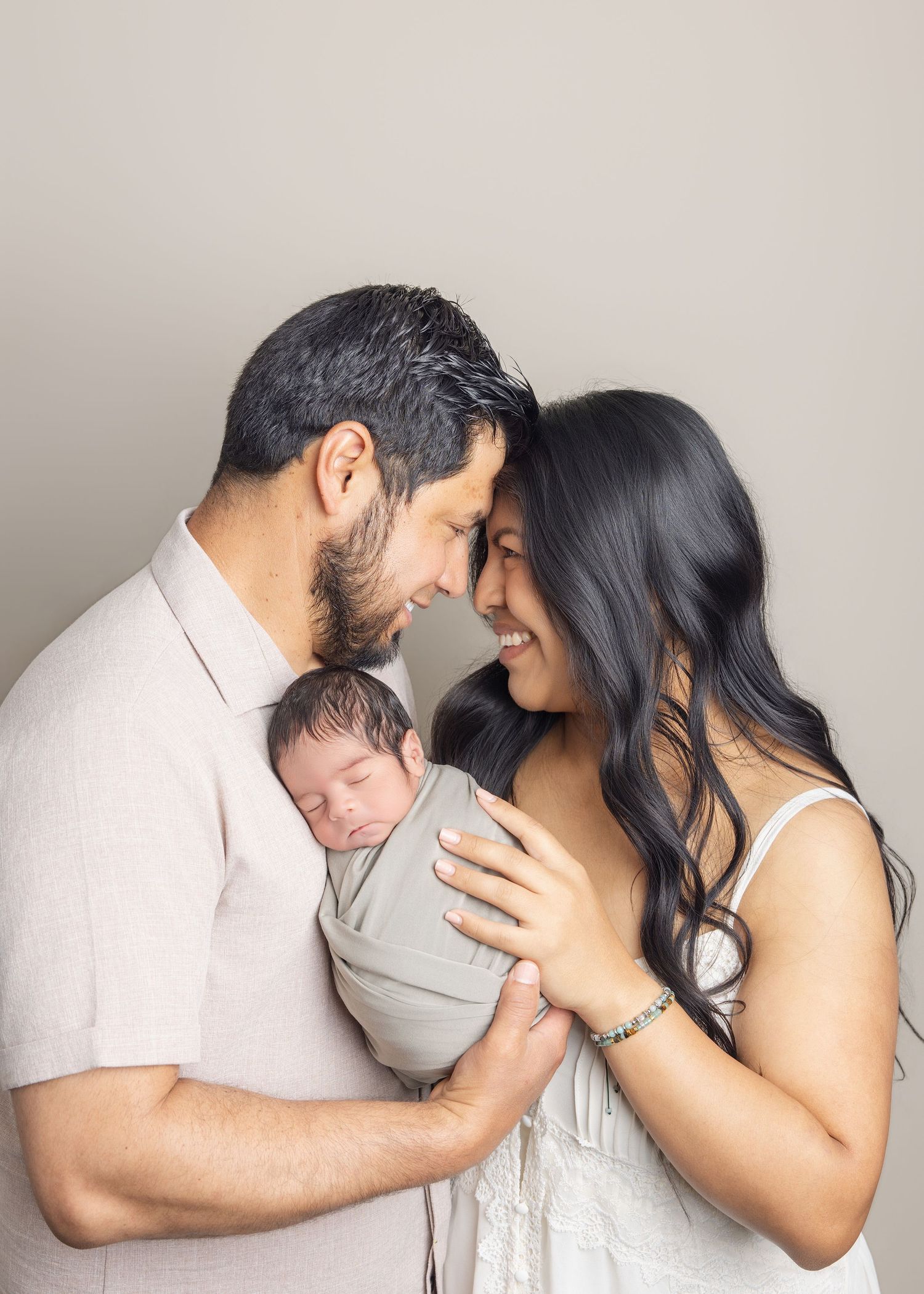 A couple tenderly touches foreheads while holding their swaddled newborn baby in a soft, neutral-toned studio portrait.