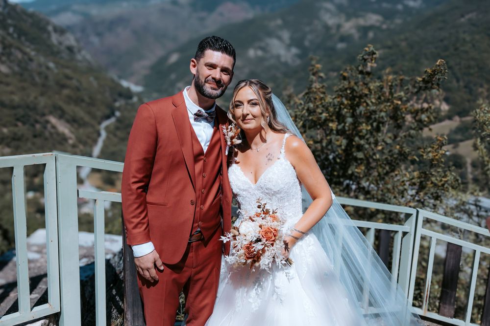 Bride and groom posing on mountain overlook, groom in rust suit, bride in white gown holding fall bouquet.
