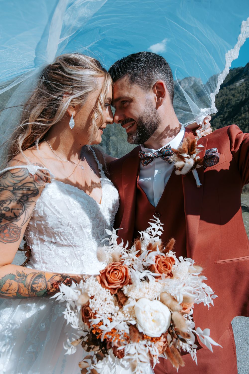 Bride and groom share an intimate moment under a veil, holding a boho bouquet of orange and white flowers outdoors.