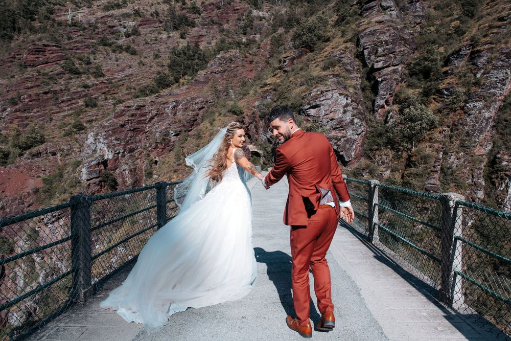 Bride in white gown and groom in red suit dance on a mountain bridge with rocky cliffs in the background.