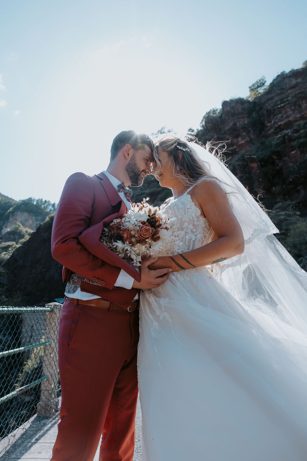 Bride and groom share romantic moment on bridge, groom in burgundy suit holding bouquet, bride in white lace gown with veil.