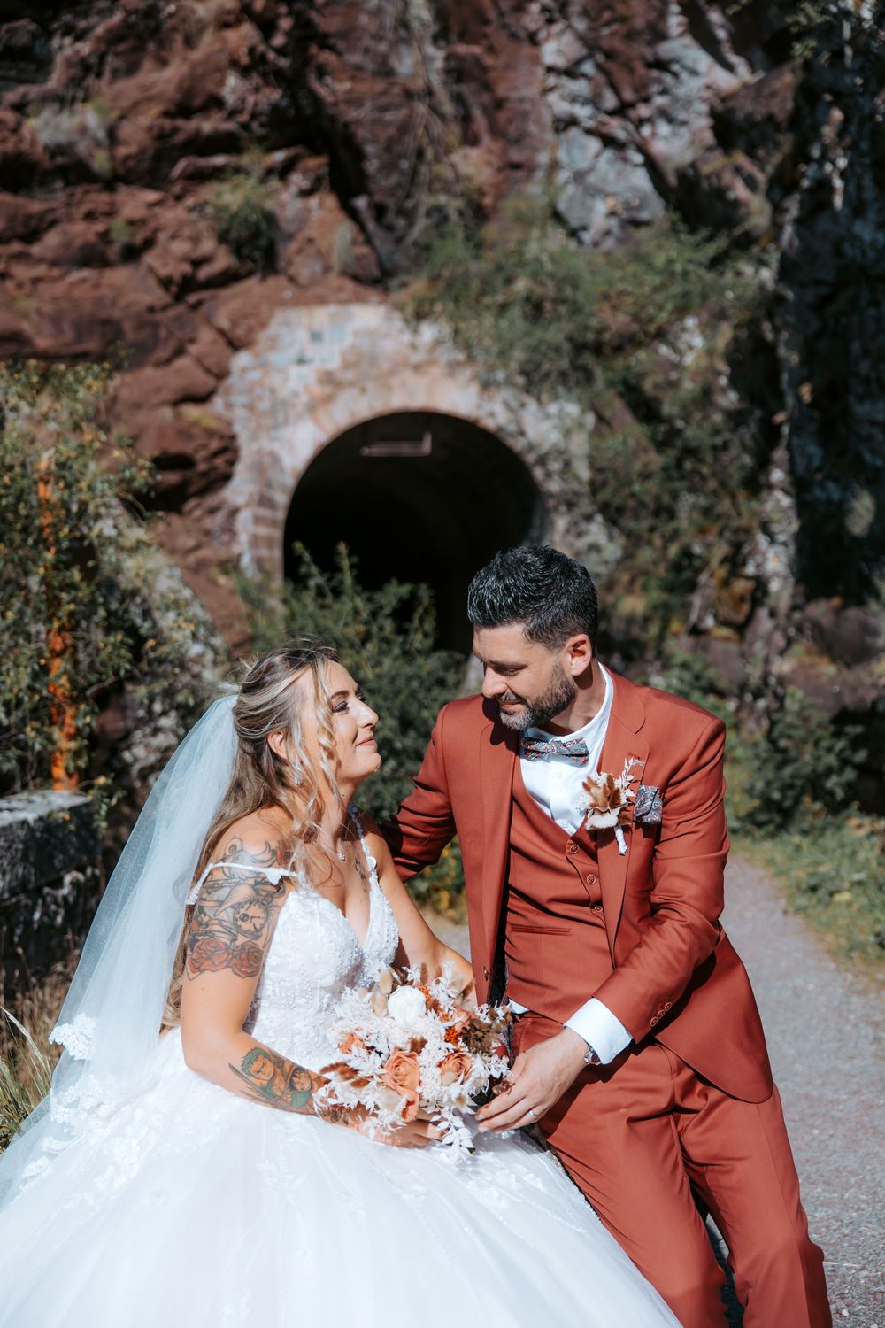 Bride and groom sitting together near a stone tunnel, bride in white gown with bouquet, groom in rust-red suit.