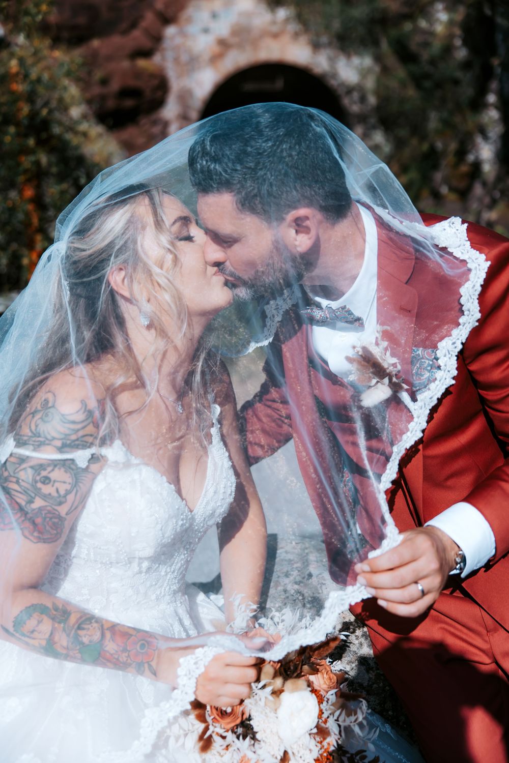 Bride and groom kissing under a wedding veil, bride wearing white dress with tattoos, groom in red suit.