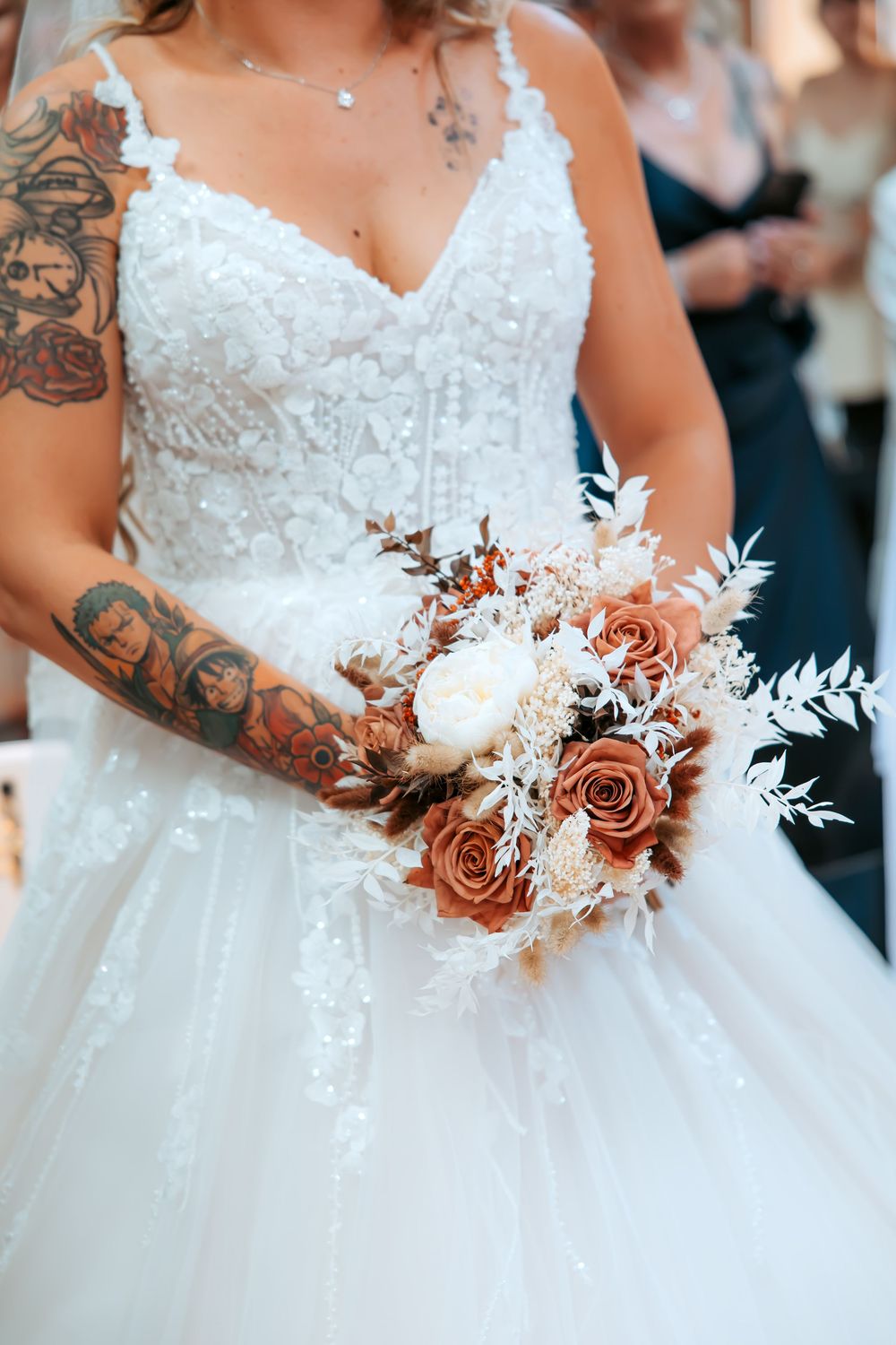 Bride in white lace gown holding a boho bouquet of terracotta roses, white peonies, and dried florals, with tattooed arms.
