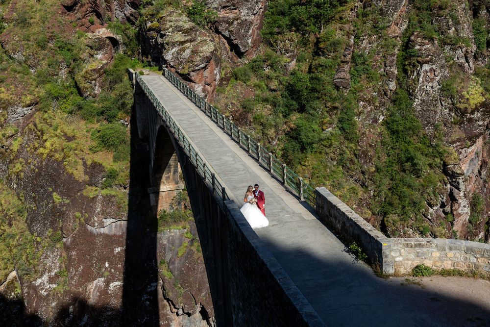 Bride and groom pose on a dramatic steel bridge over a rocky gorge surrounded by lush green cliffs.