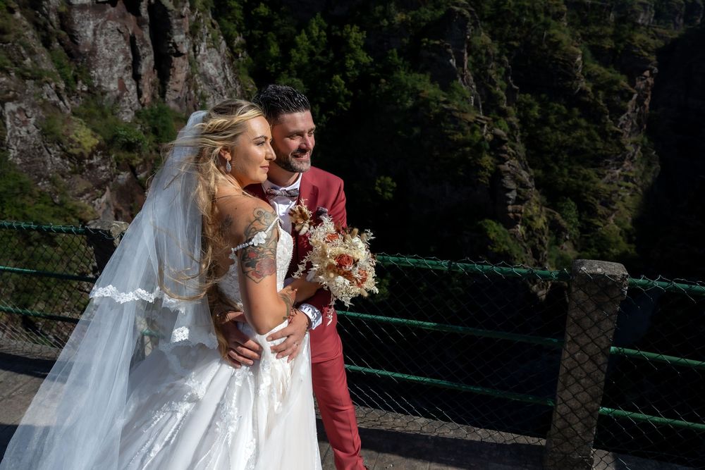 Bride in white gown and groom in red suit pose on a bridge with dramatic mountain cliffs in background.