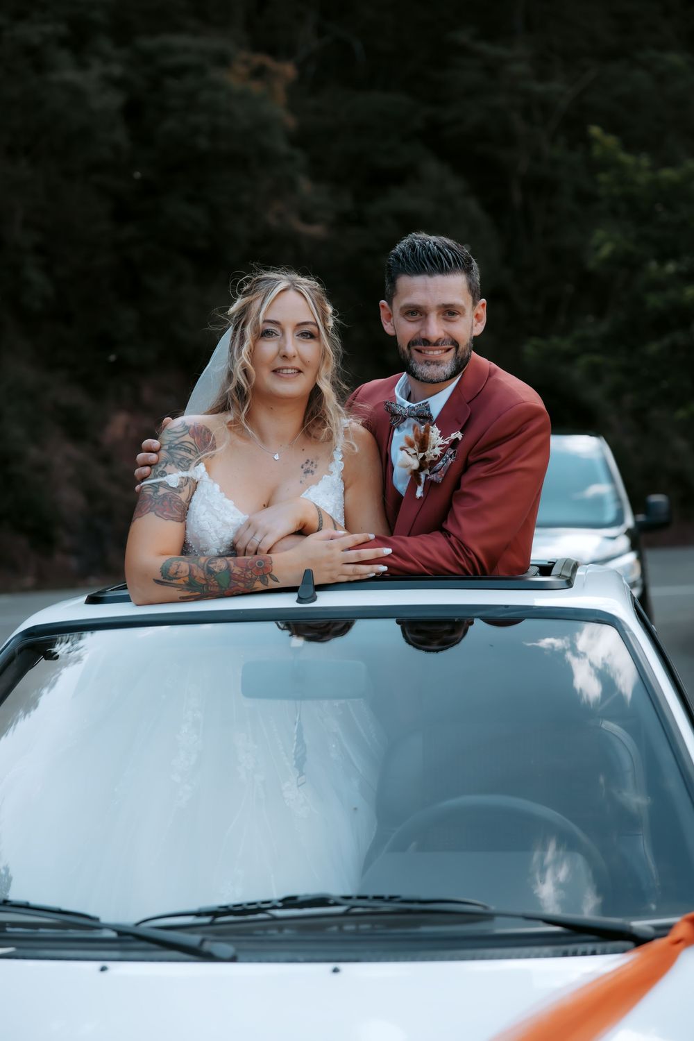Bride and groom smiling through sunroof of white car, bride in veil, groom in burgundy suit with boutonniere.