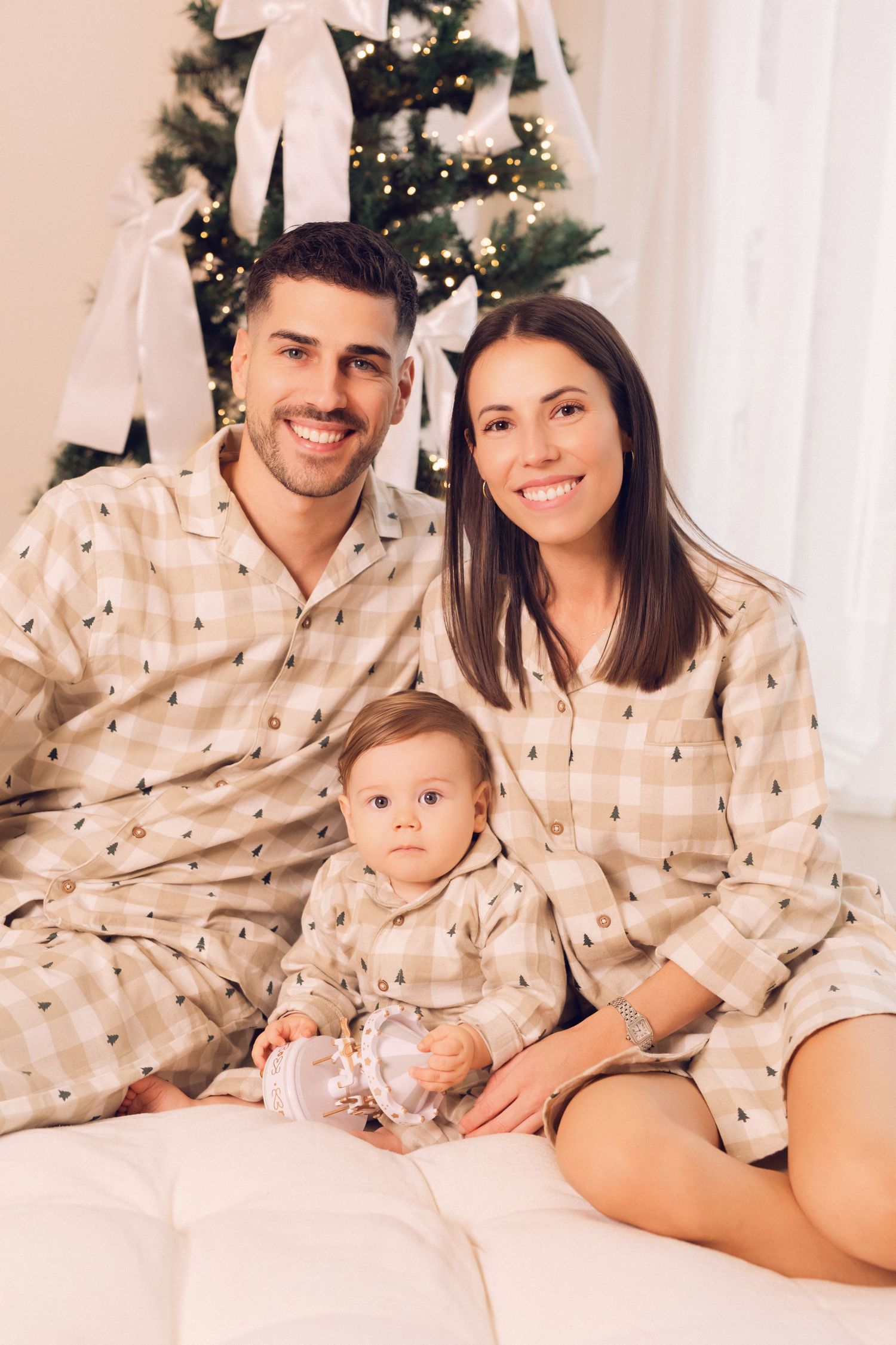 Happy family in matching plaid Christmas pajamas sitting together near a decorated holiday tree with a baby.